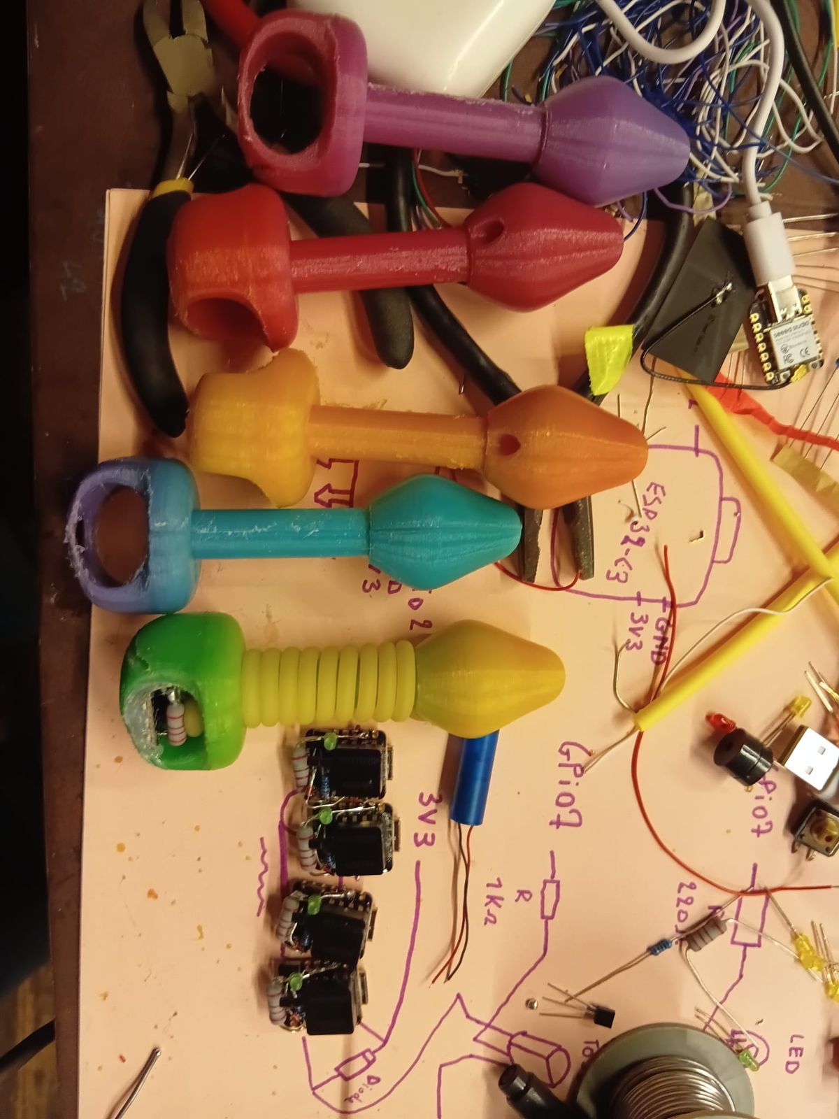 Printed plugs in several colours on a workbench, among pliers, wires, an ESP32-C3 board and a hand-drawn wiring sketch.